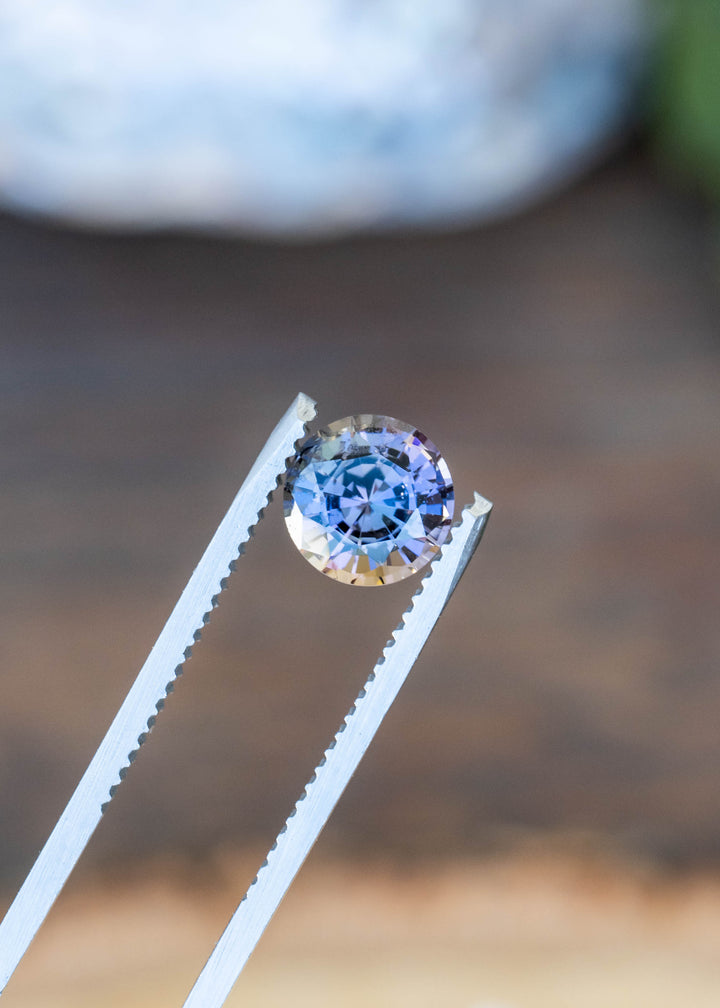 Close-up of a natural 1.72 carat untreated custom round cut tanzanite in tweezers highlighting its exceptional blue, purple, green, and amber pleochroism.
