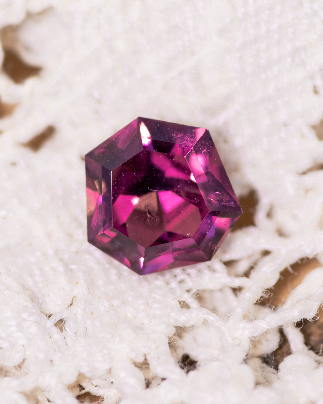 1.00 carat red pink purple garnet resting on a soft lace fabric, showing vivid brilliance and the unique symmetry of its mix geometric shield cut, untreated gemstone sourced from Kenya.