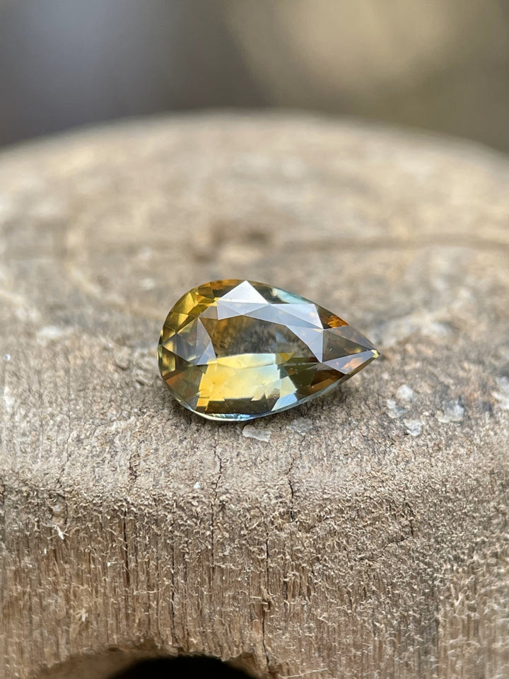 A 1.00-carat mixed pear brilliant sapphire resting on a rustic wood surface, displaying unique bi-color zoning in brown and bluish green.