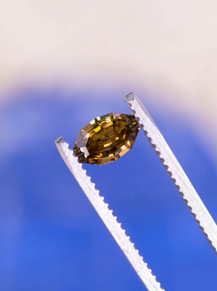 Loose fancy orangish yellow brown diamond held in tweezers displaying the elongated moval shape and antique step cut facet pattern.
