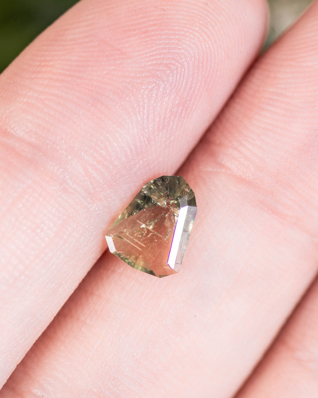 1.01 carat yellow, teal, and green sapphire resting on fingertip, showcasing natural brilliance and geometric elegance of the custom shield portrait cut, untreated sapphire from Umba River, Tanzania.