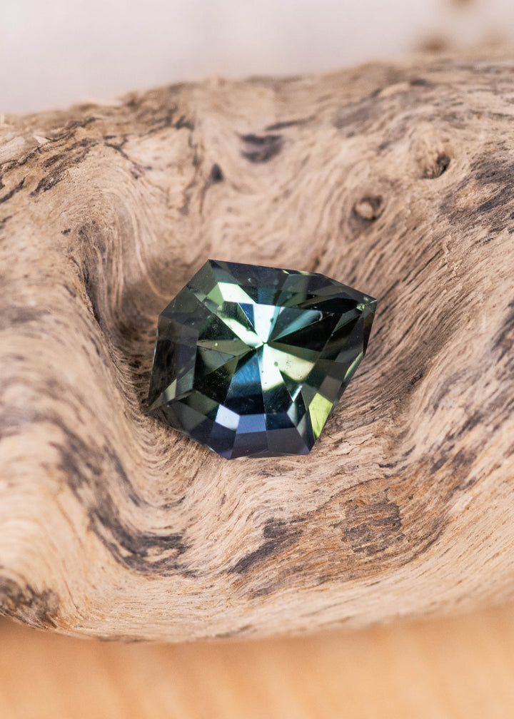 Loose teal blue green tanzanite resting on natural wood showing the custom geometric shield cut and color shifting facets from Merelani Hills Tanzania.