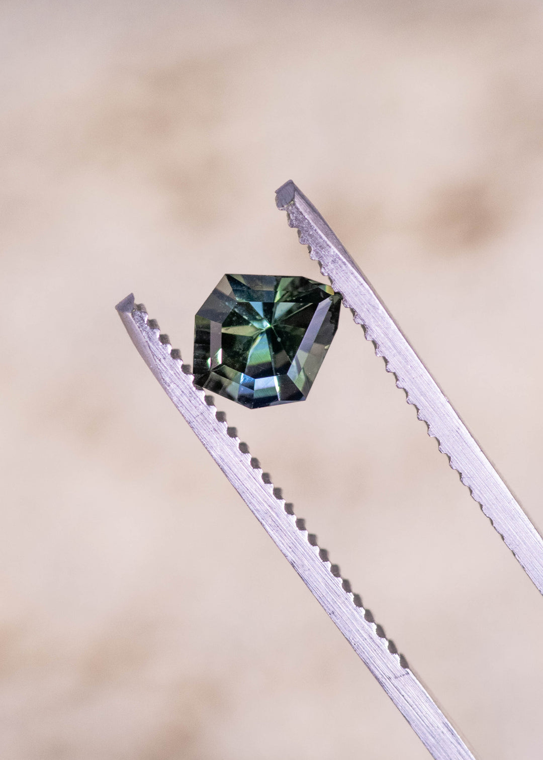 Teal blue green tanzanite gemstone held in tweezers displaying the precision geometric mix cut shield shape and reflective teal blue flashes.
