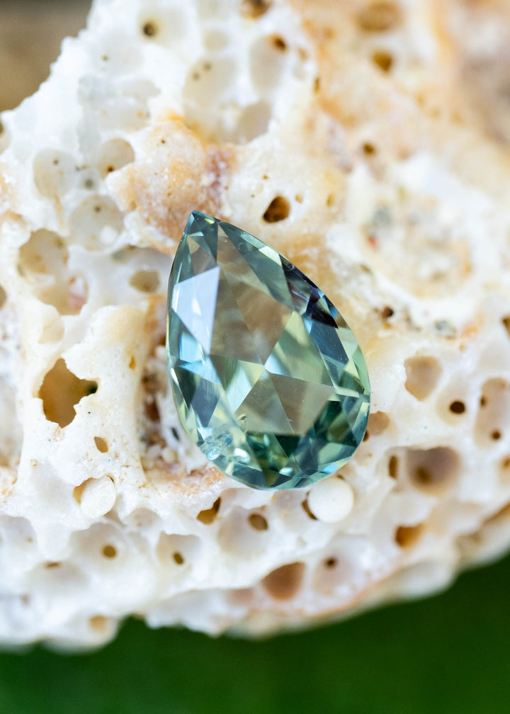 Pear-shaped green sapphire resting on textured white coral, with teal-blue flashes along its facets and deeper green color near the rounded end.
