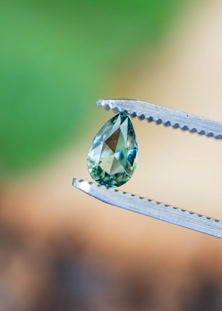Small pear-shaped sapphire held upright between silver tweezers against a blurred green and beige background, showing its pointed tip and green-to-teal faceting.