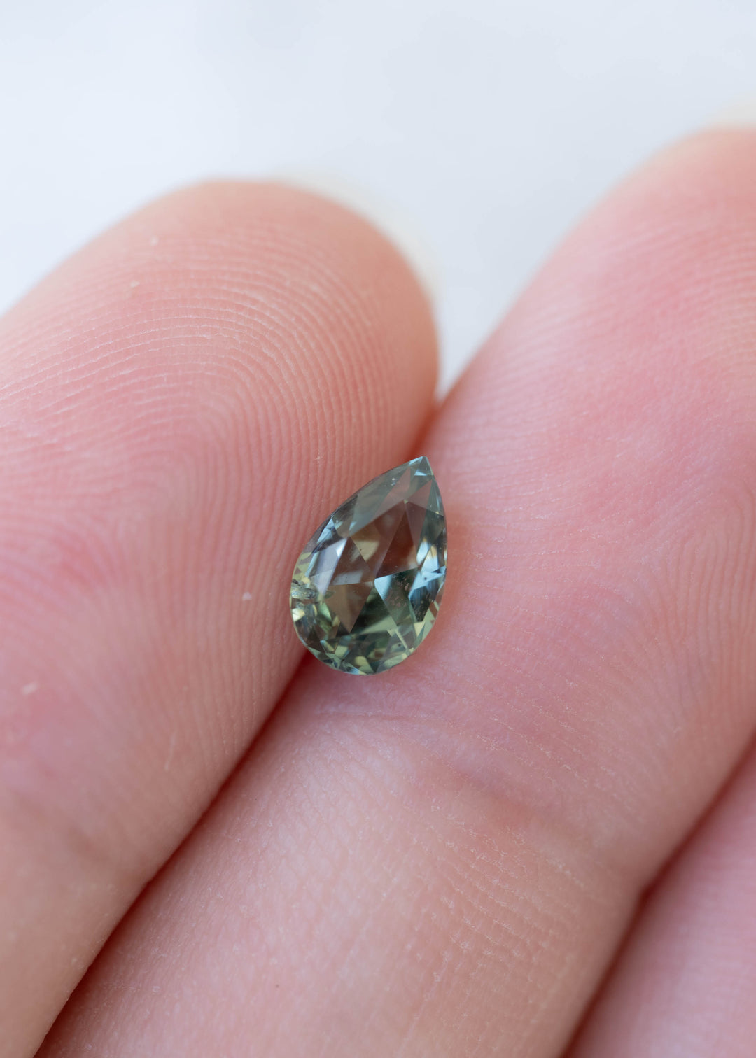 Loose pear-shaped green sapphire resting between two fingertips, displaying a pointed tip, rounded base and muted teal and forest green facets.