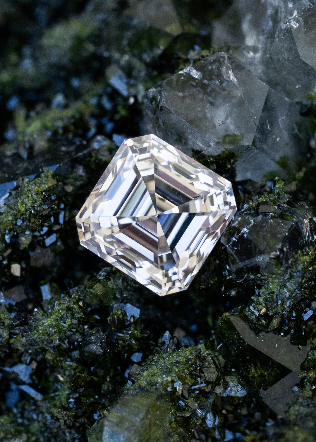 1.02 carat M color VVS1 Asscher cut Canadian diamond resting among dark green mineral crystals, showing bold clipped corners and concentric geometric step facets.