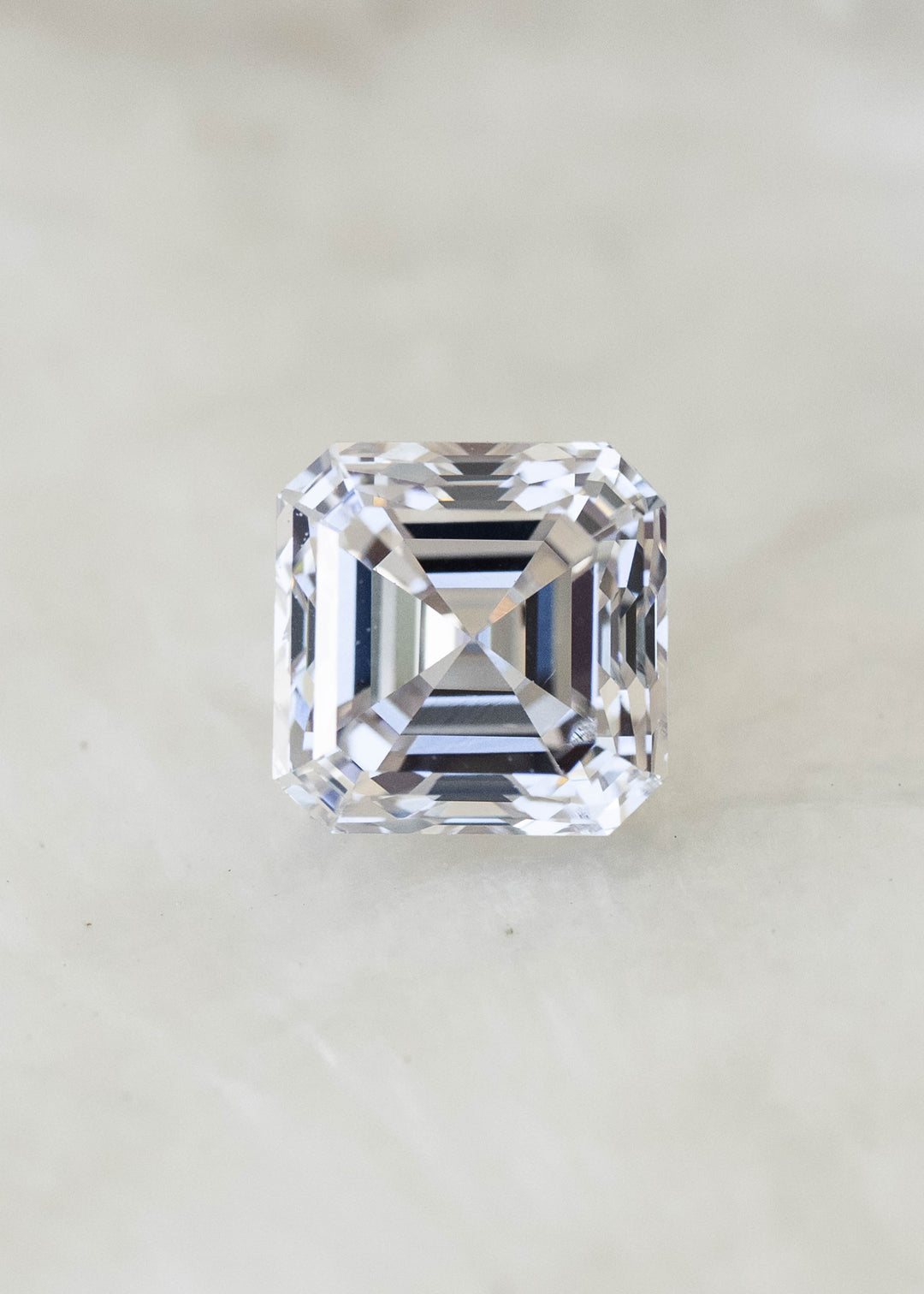 Face-up macro photograph of a 1.02 carat natural Asscher cut diamond on a pale background, showing its nearly square proportions and crisp hall-of-mirrors facet pattern.