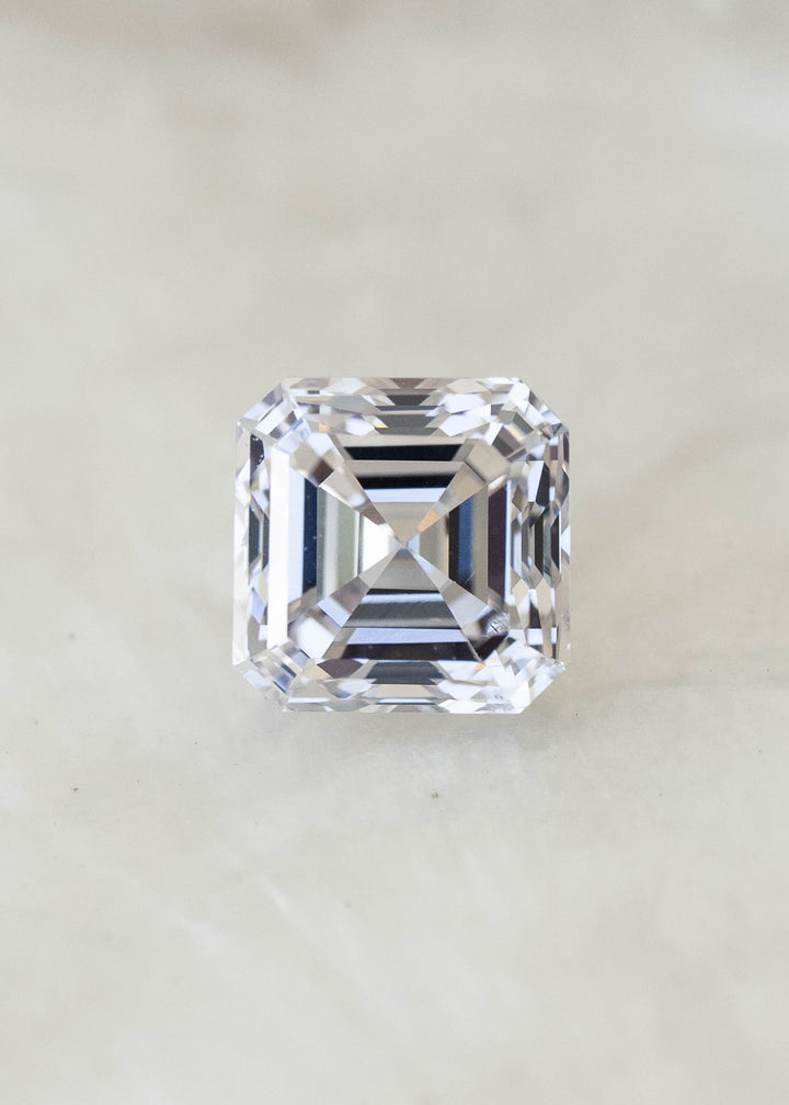 Face-up macro photograph of a 1.02 carat natural Asscher cut diamond on a pale background, showing its nearly square proportions and crisp hall-of-mirrors facet pattern.