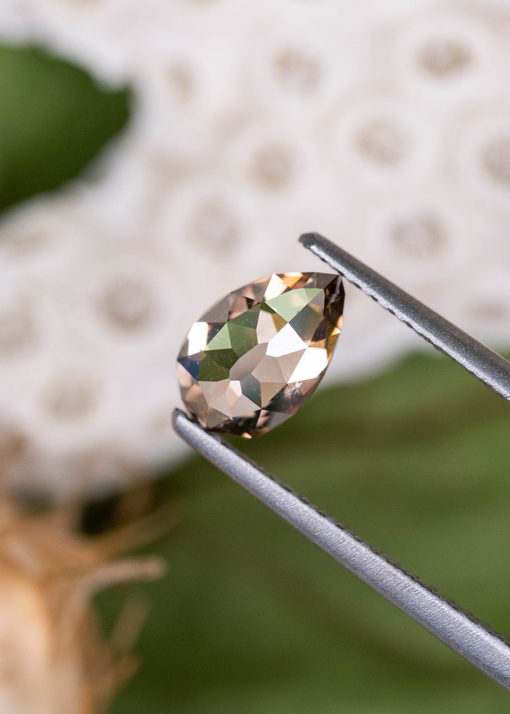 Close-up of a 1.02ct South African Fancy Orangish Brown diamond held in jeweler's tweezers. Its geometric Mughal faceting reveals contrasting reflections of amber, smoky brown, and silvery white, emphasizing the unusual pear-shaped cut.