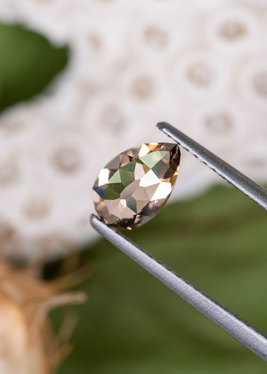 Close-up of a 1.02ct South African Fancy Orangish Brown diamond held in jeweler's tweezers. Its geometric Mughal faceting reveals contrasting reflections of amber, smoky brown, and silvery white, emphasizing the unusual pear-shaped cut.