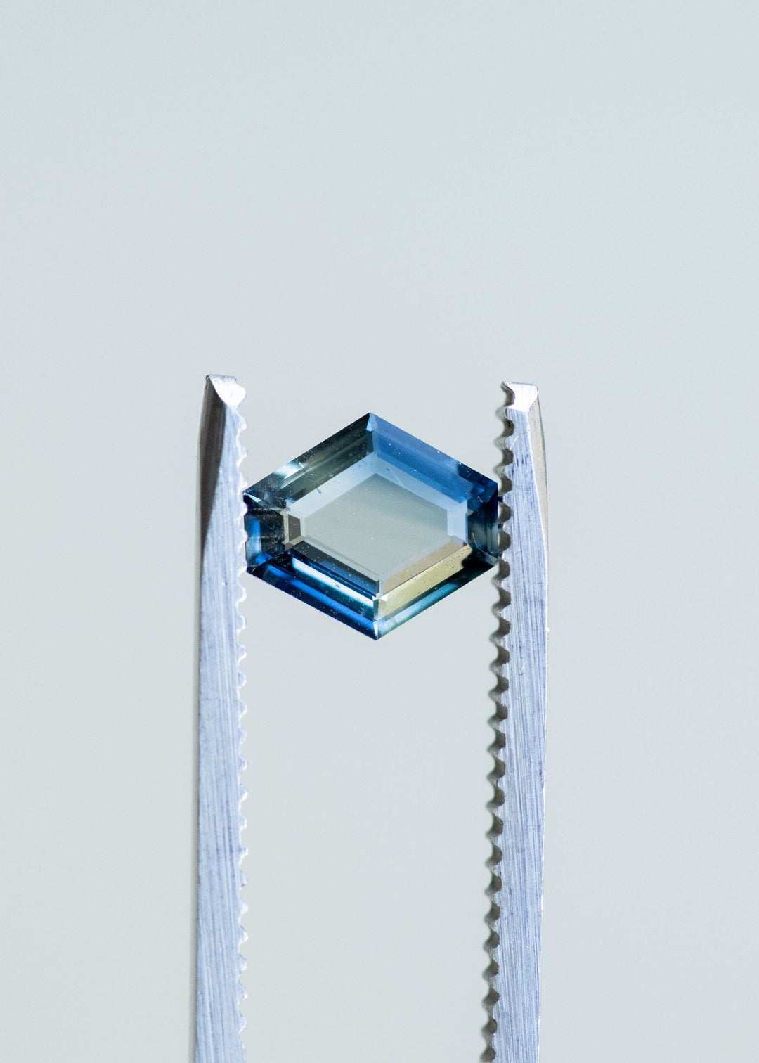 Hexagonal portrait cut teal blue 1.02ct sapphire held in gem tweezers in natural light showcasing natural parti color zoning.