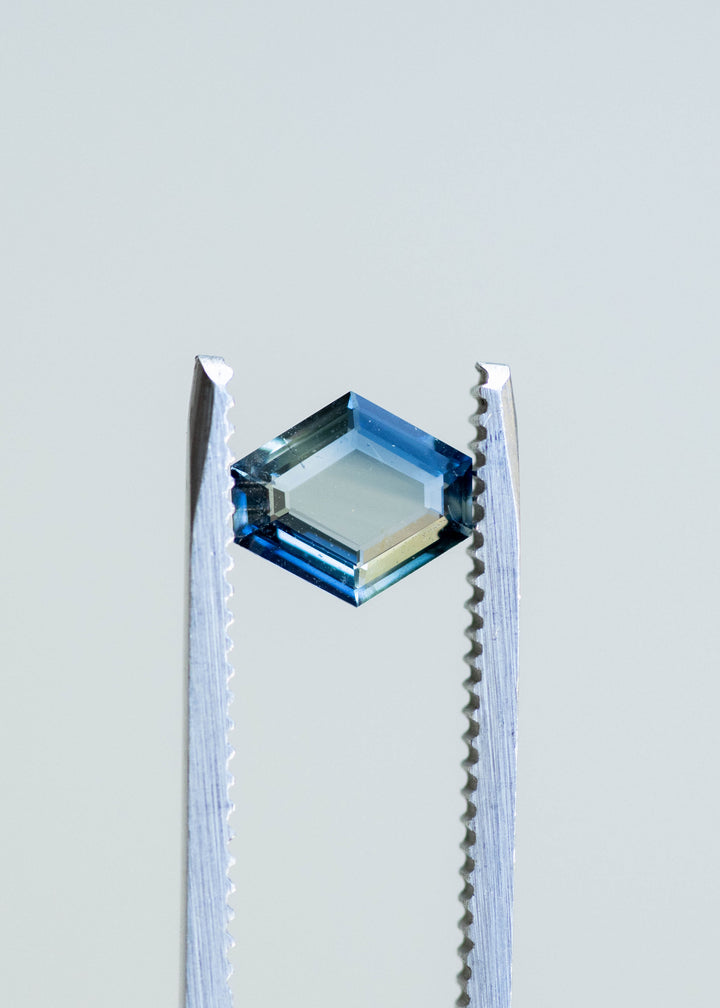 Hexagonal portrait cut teal blue 1.02ct sapphire held in gem tweezers in natural light showcasing natural parti color zoning.
