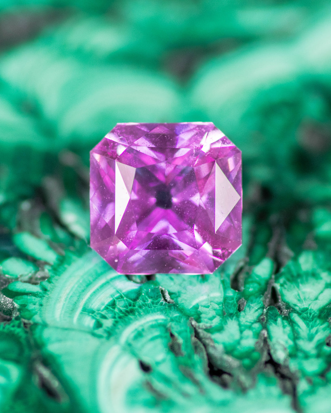 1.03 carat pink and purple sapphire resting on textured green malachite background, highlighting vivid color contrast and brilliance of its square scissor cut, untreated sapphire from Sakaraha, Madagascar.