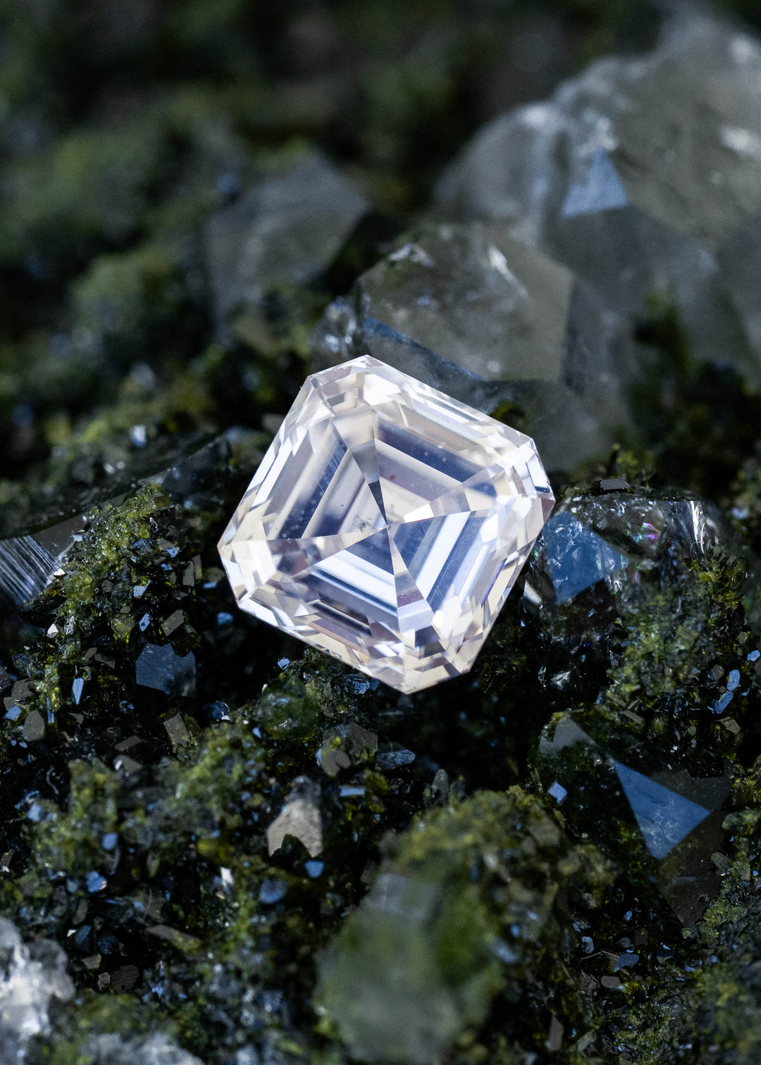 1.03 carat M color VVS1 Asscher cut Canadian diamond resting among dark green mineral crystals, photographed at an angle to show its clipped corners and layered step facets.