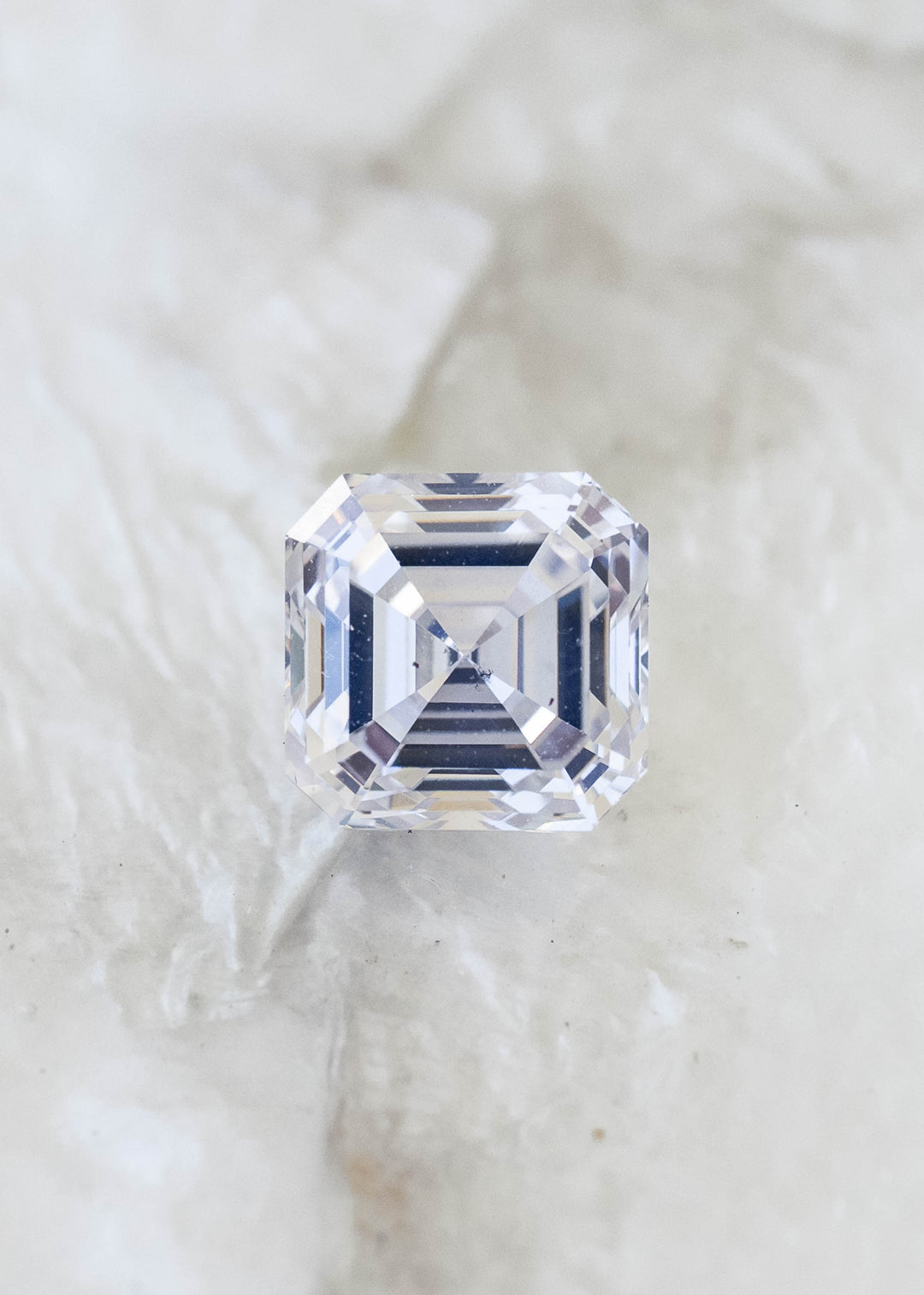 Face-up macro view of a 1.03 carat natural Asscher cut diamond on a pale mineral background, showing concentric step facets and subtle ivory warmth.