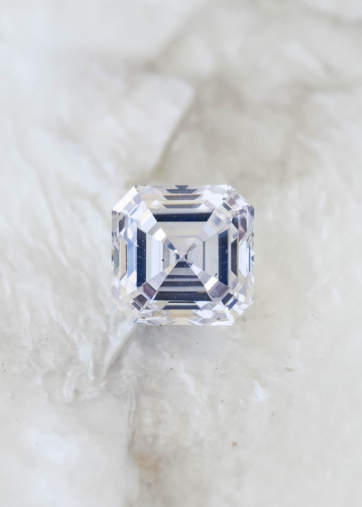 Face-up macro view of a 1.03 carat natural Asscher cut diamond on a pale mineral background, showing concentric step facets and subtle ivory warmth.