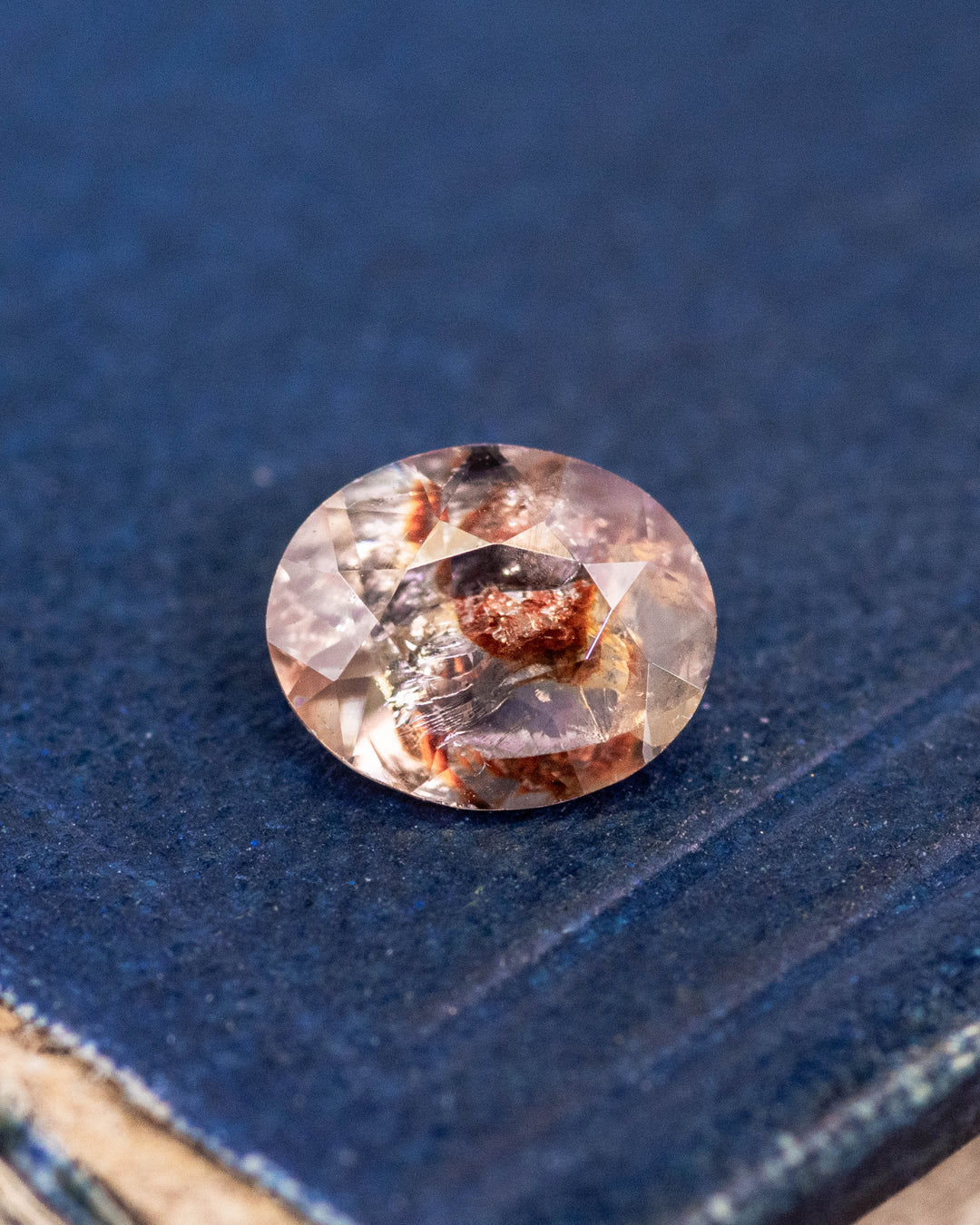 1.05 carat orange, peach, and red sapphire resting on textured blue surface, showing natural inclusions and brilliance of custom oval cut, untreated gemstone from Umba River Valley, Tanga, Tanzania.