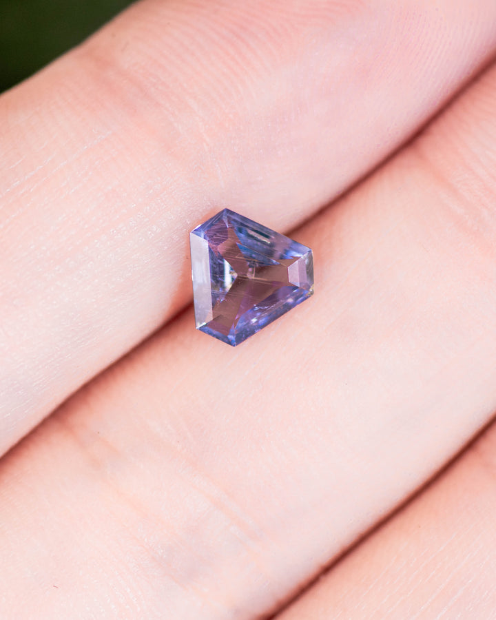 1.05 carat violet, aqua, and blue sapphire displayed on fingertip under natural light, showing unique custom cut corner trillion shape and natural untreated clarity, sourced from Umba River, Tanzania.