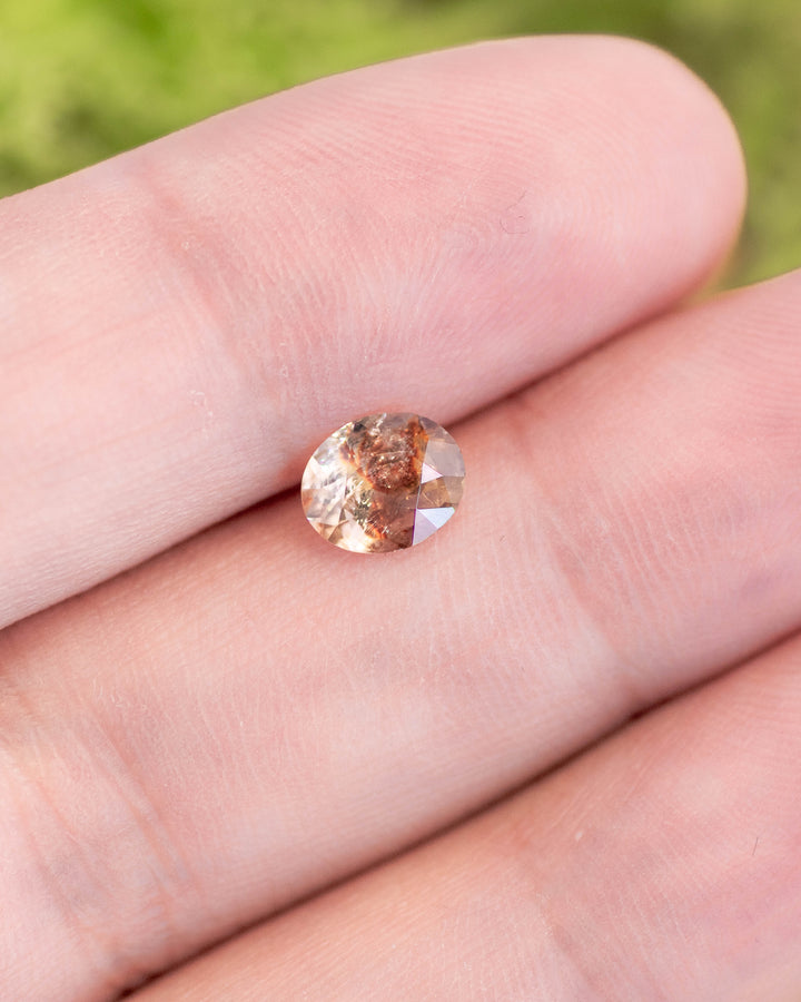 1.05 carat orange, peach, and red sapphire displayed on fingertip under natural light, highlighting the earthy inclusions and fire of its custom oval cut, untreated gemstone from Umba River Valley, Tanzania.