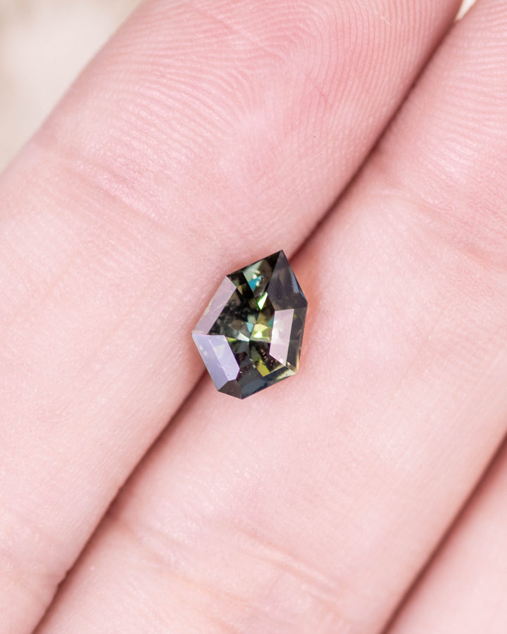 1.05 carat tanzanite displayed on fingertip under natural light, showing the interplay of yellow, green, and blue hues with sharp geometric shield facets, untreated gemstone from Merelani Hills, Arusha Region, Tanzania.