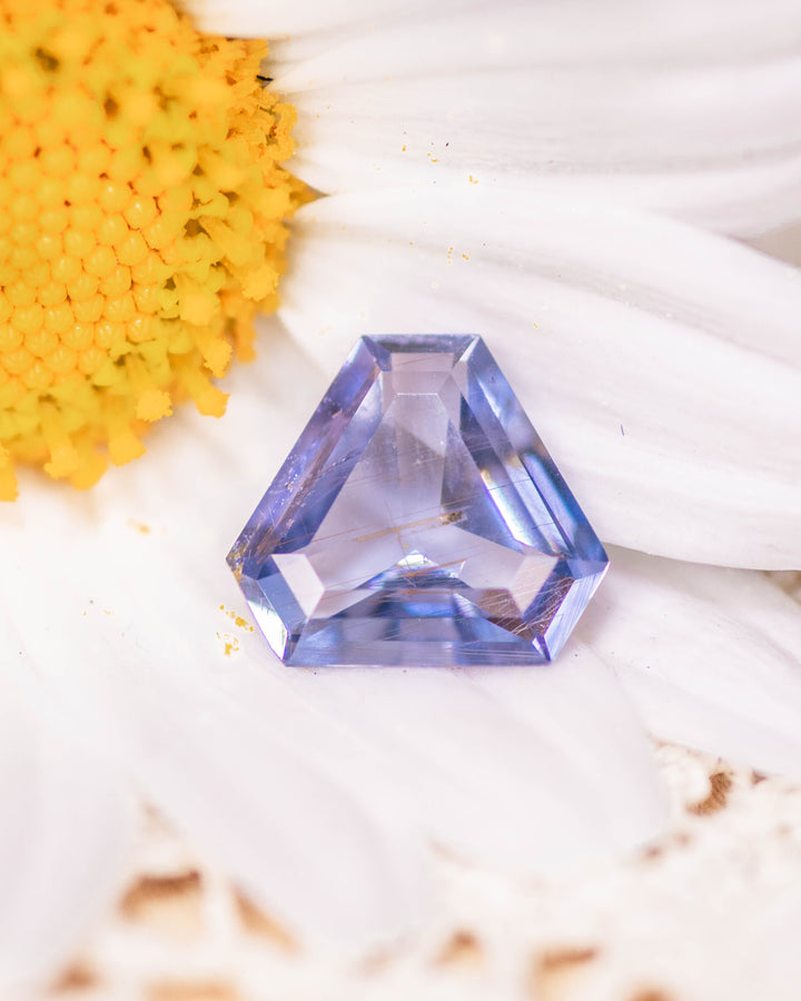 1.05 carat violet, aqua, and blue sapphire resting on white flower petals, showcasing its custom cut corner trillion mix cut brilliance, natural untreated sapphire from Umba River, Tanzania.