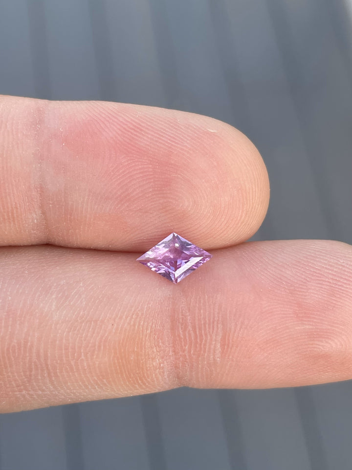Close-up of a 1.06-carat purple sapphire from Sri Lanka in a unique kite step cut, resting on a fingertip to showcase its elegant color and clarity. The gemstone features a soft purple hue with exceptional precision, making it ideal for fine jewelry.