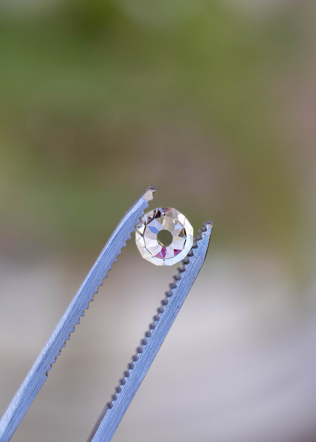 Round Mughal cut South African diamond held face-up in jeweler’s tweezers against a softly blurred natural background, showing its open center and floral-like facet arrangement.