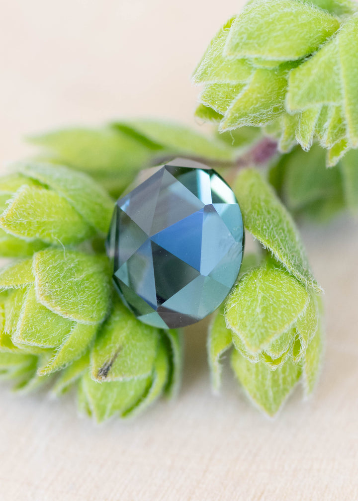 Teal blue green oval double rose cut sapphire resting between green botanical leaves
