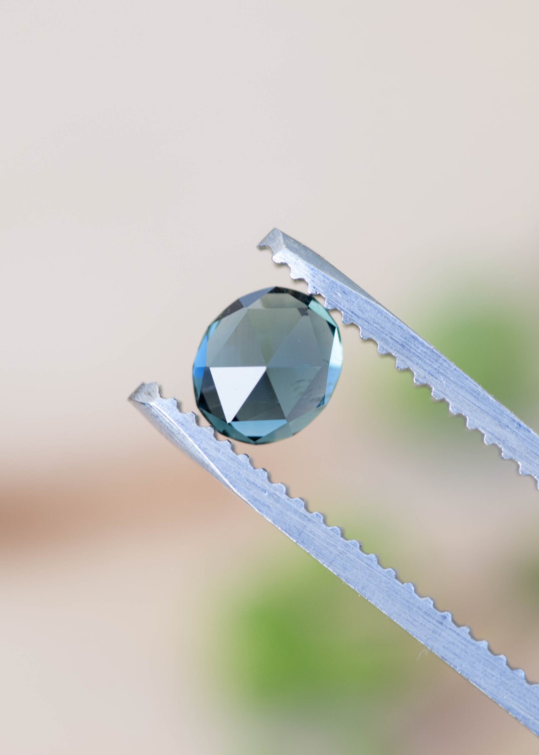 Close-up of teal blue green oval rose cut sapphire showing geometric faceting held by tweezers against a blurred background