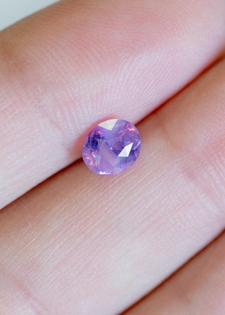 Loose opalescent purple pink sapphire on fingertip, revealing balanced oval proportions and delicate violet-to-blush color shift in natural light.