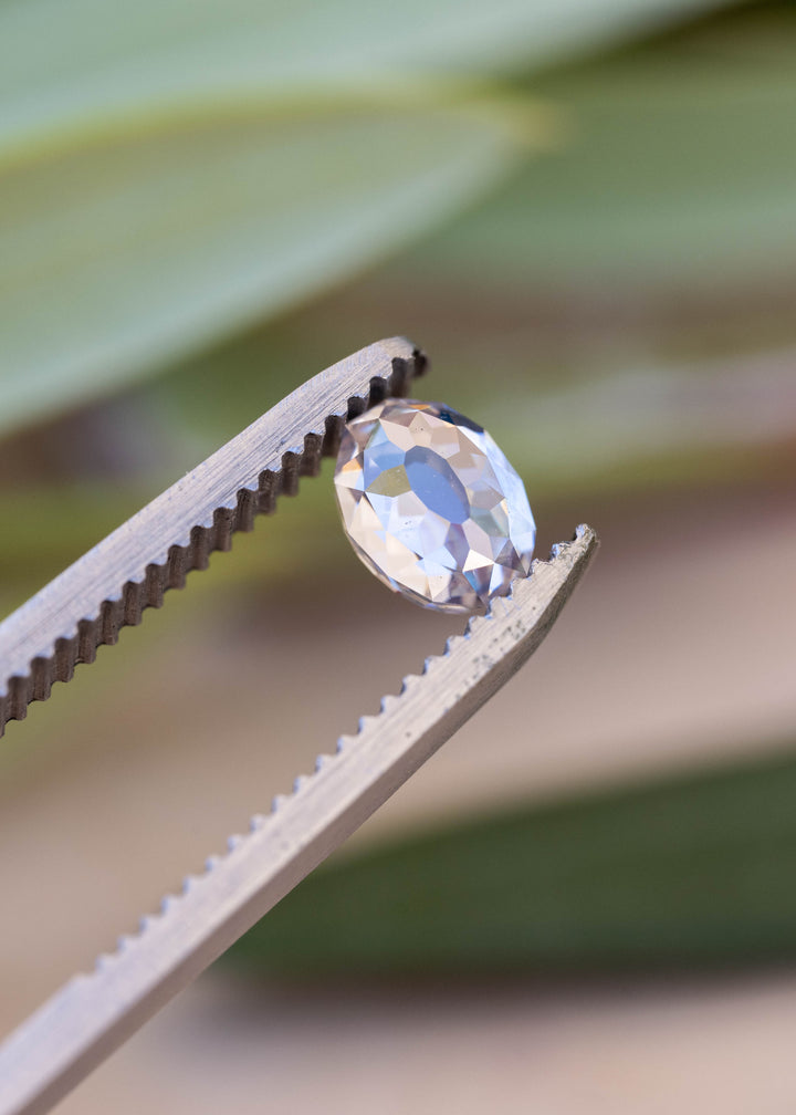 Oval Mughal cut diamond held between jeweler’s tweezers against a softly blurred botanical background, displaying pale champagne, white and blue reflections across its broad facets.
