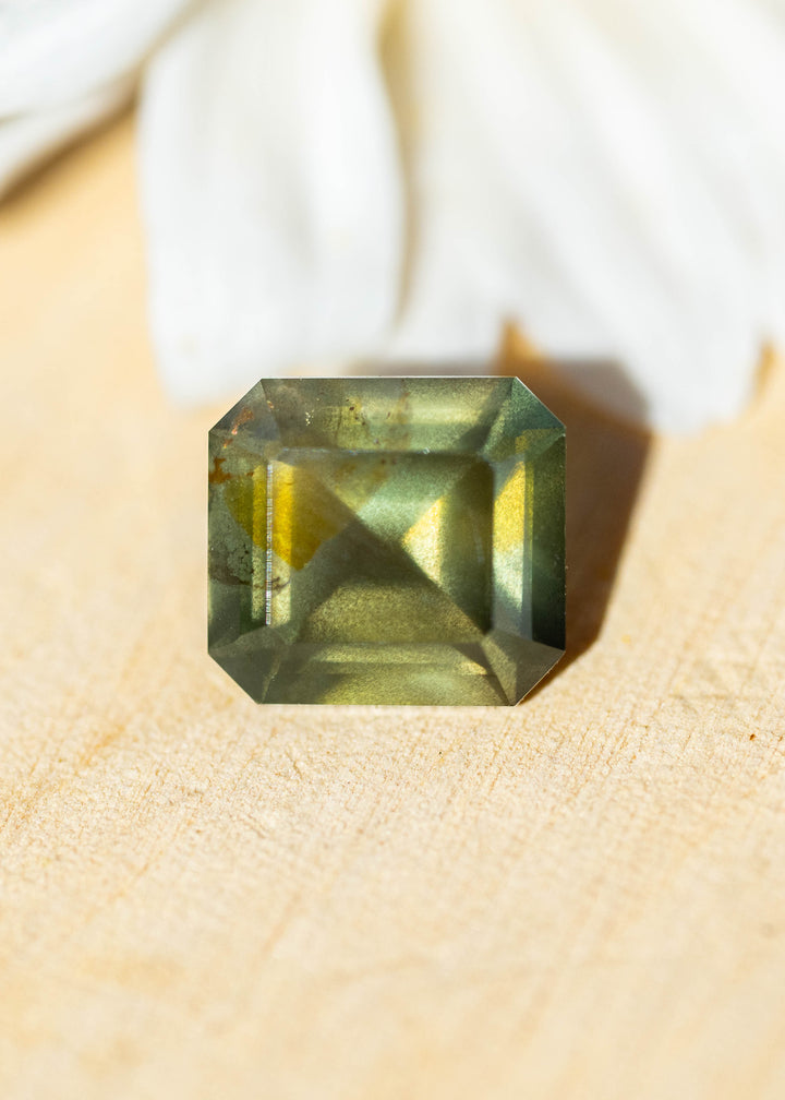 Natural 1.08 carat opalescent moss green sapphire from Gombe, Nigeria, photographed on a light natural surface. Its precision octagonal step mix cut reveals olive-green coloration, golden reflections and visible internal texture.