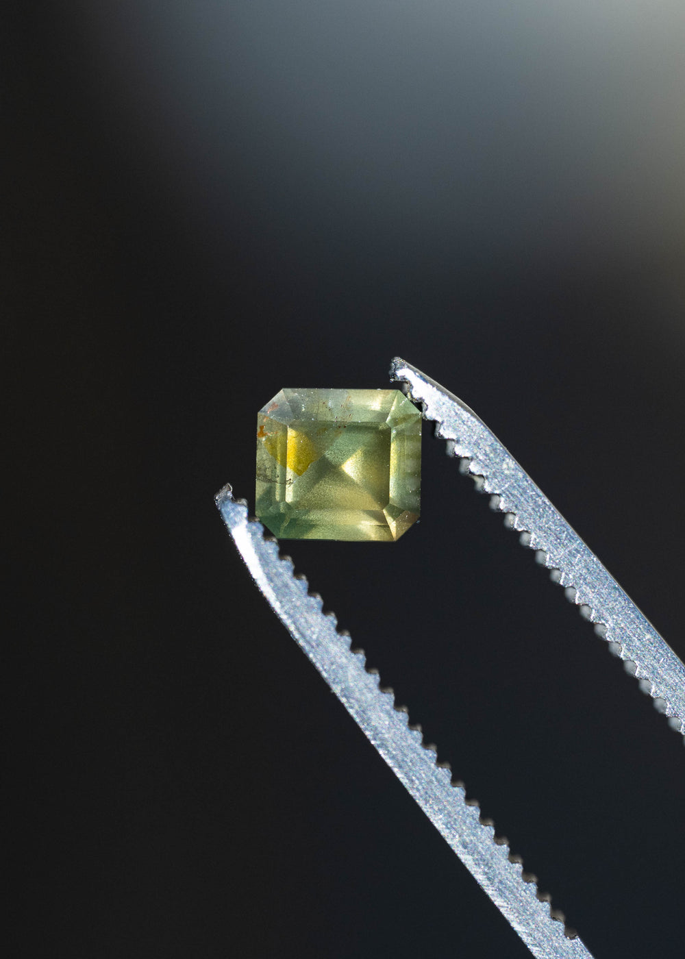 Close-up of an untreated 1.08ct moss green sapphire held in silver jeweler's tweezers, highlighting its clipped-corner octagonal outline, step-style facets and muted olive-green color.
