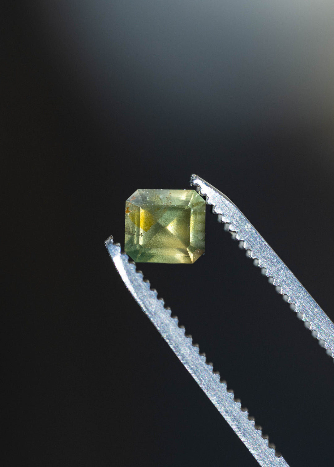 Close-up of an untreated 1.08ct moss green sapphire held in silver jeweler's tweezers, highlighting its clipped-corner octagonal outline, step-style facets and muted olive-green color.