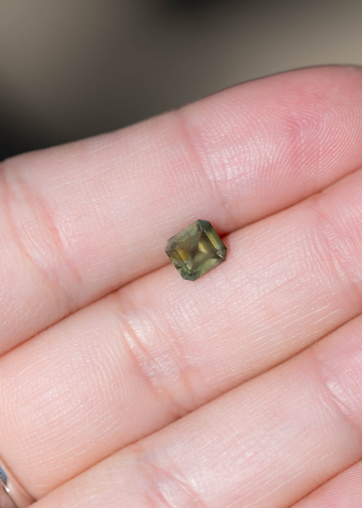 Untreated 1.08 carat opalescent moss green sapphire from Nigeria photographed on fingers for scale. Its precision octagonal step mix cut measures 5.56 × 5.11 mm.