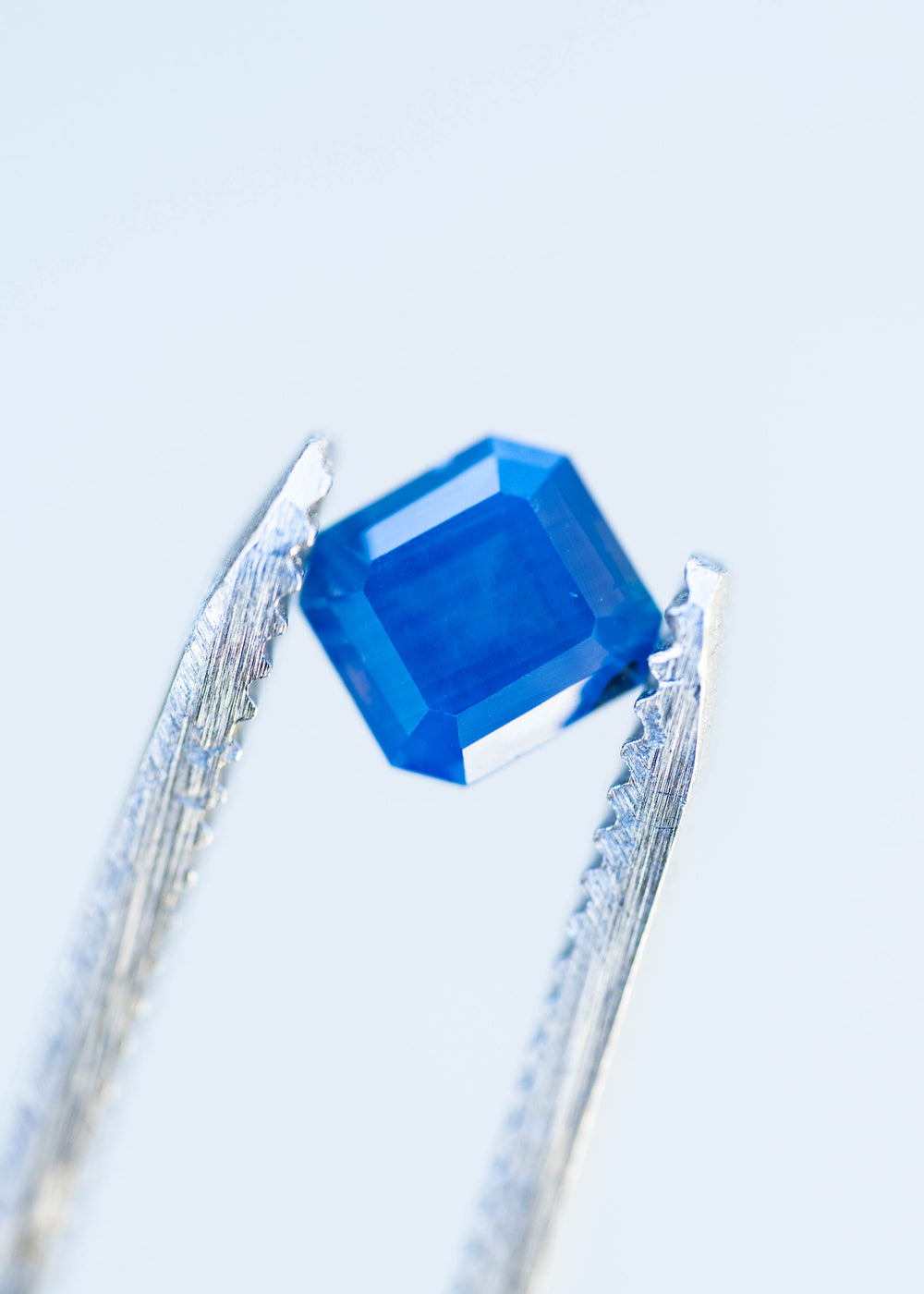 Close-up of a 1.08ct untreated Nigerian sapphire held between jeweler's tweezers. Its squared octagonal step cut features clipped corners, broad geometric facets and saturated blue coloration.