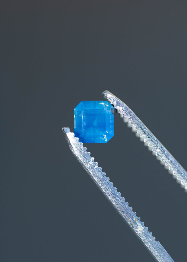 Detailed photograph of an untreated opalescent denim blue sapphire from Gombe, Nigeria. The stone's compact octagonal outline and rich blue color are shown against a dark gray background.