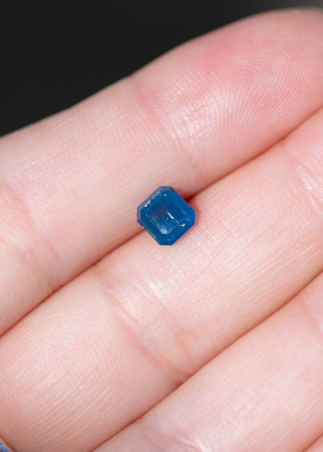 Natural 1.08 carat opalescent denim blue sapphire photographed on fingers for scale. Its squared octagonal step cut measures 5.61 × 5.30 mm face-up and features a deep blue center.