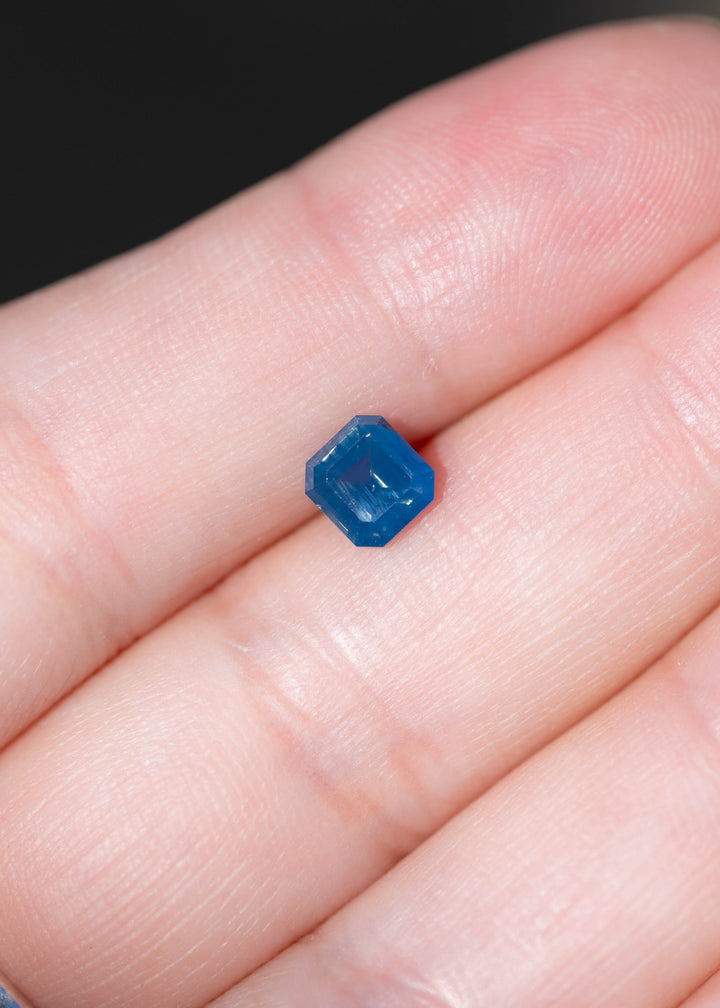 Natural 1.08 carat opalescent denim blue sapphire photographed on fingers for scale. Its squared octagonal step cut measures 5.61 × 5.30 mm face-up and features a deep blue center.