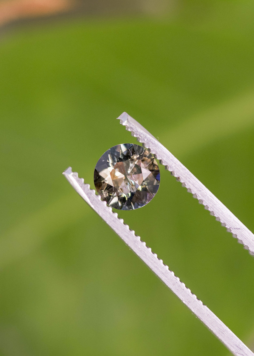 1.08ct blue and brown tanzanite custom modified round brilliant held in gem tweezers, highlighting sparkle, internal inclusions and rainbows, and natural untreated gemstone from Merelani Hills Tanzania.