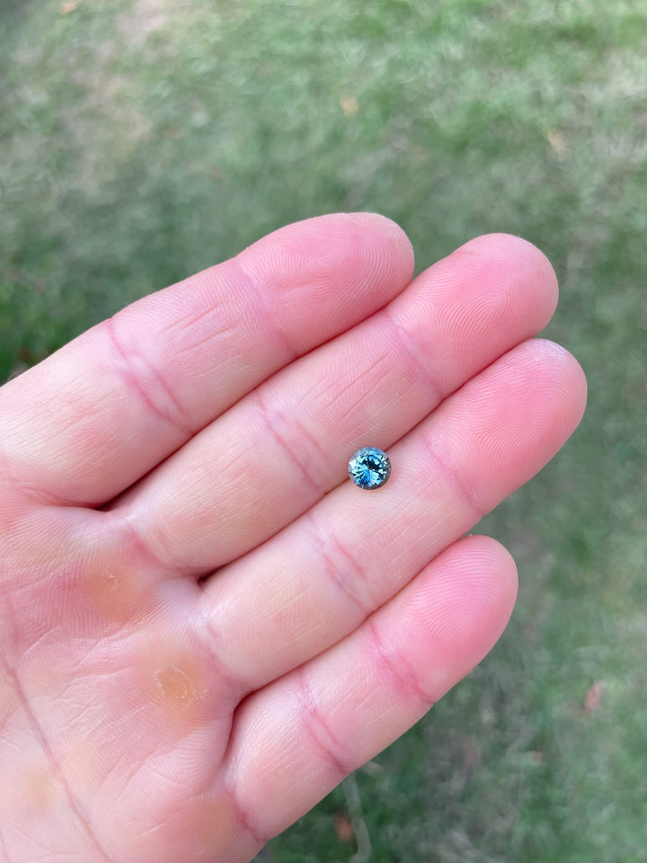 A 1.08-carat round brilliant sapphire from Madagascar, featuring a mix of teal, blue, and green hues, shown on an open palm in natural light.