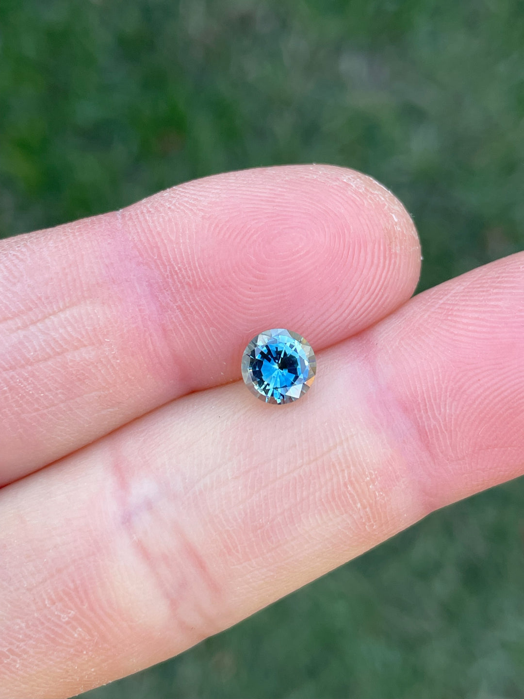 A close-up of a 1.08-carat round brilliant sapphire balanced on a fingertip, with deep teal and blue-green colors sparkling under daylight.