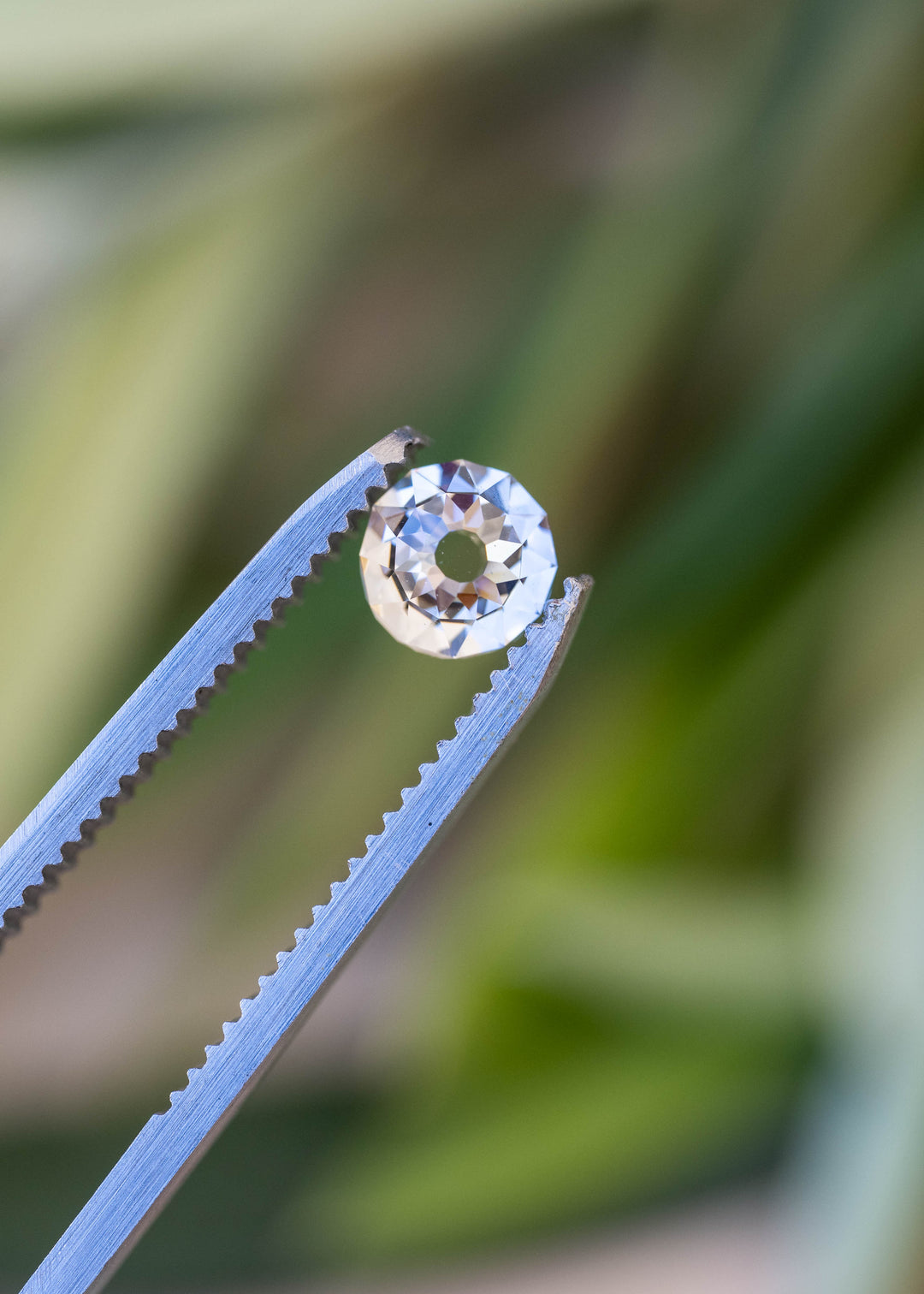 Round Mughal cut South African diamond held between jeweler’s tweezers against a softly blurred green background, showing the open center and surrounding geometric facets.