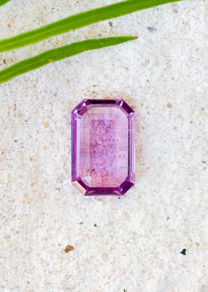 Natural pink sapphire in portrait cut resting on textured white surface with foliage in the corner.