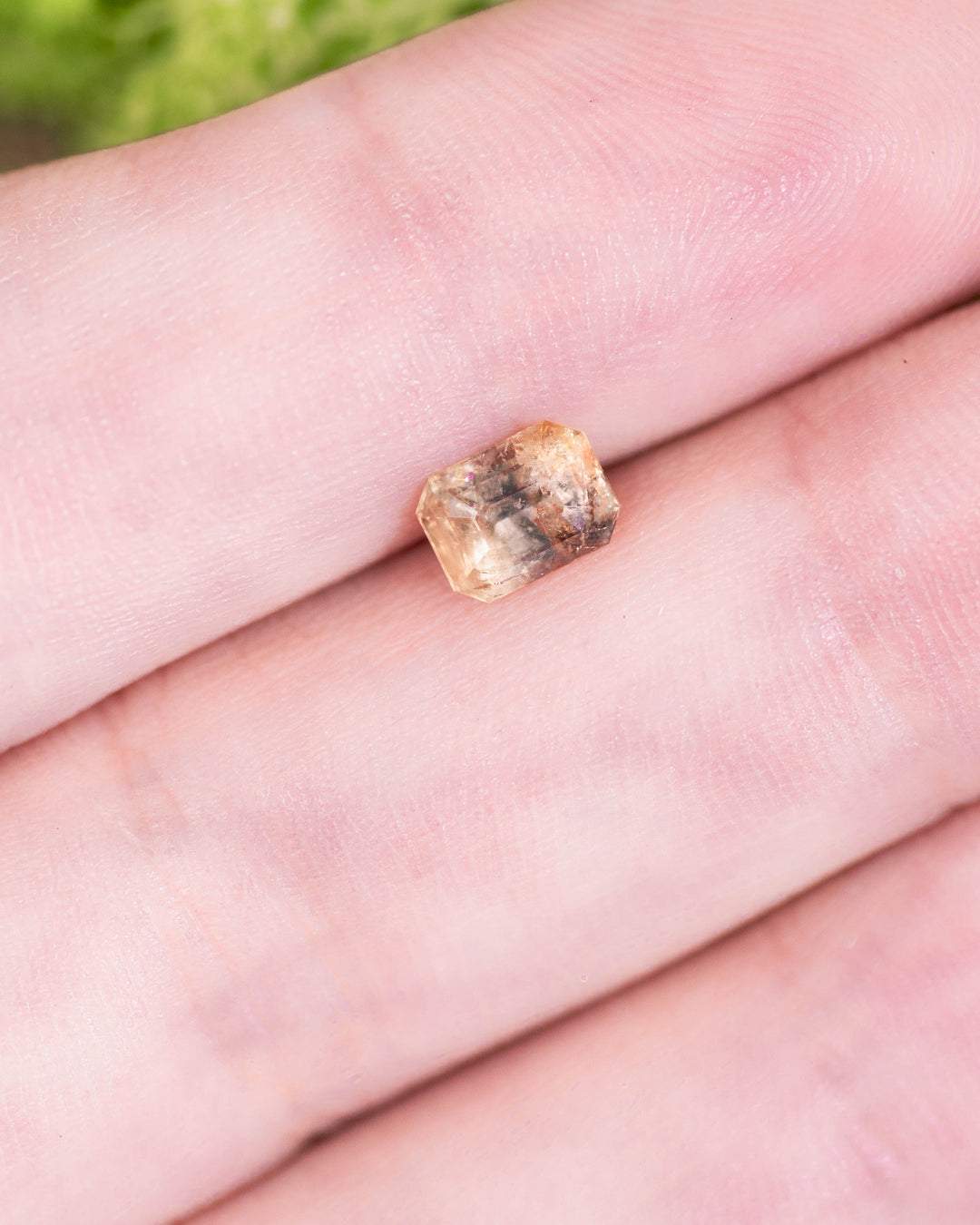 1.11 carat grey, teal, and yellow parti sapphire resting on a fingertip, showcasing its unique custom emerald cut and natural inclusions, untreated gemstone from Umba River Valley, Tanga, Tanzania.