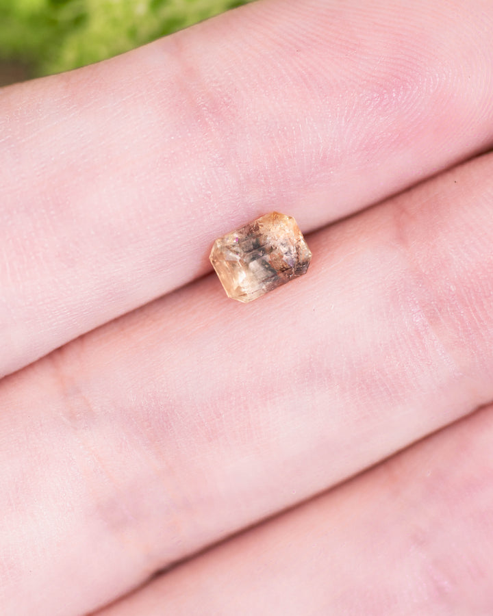 1.11 carat grey, teal, and yellow parti sapphire resting on a fingertip, showcasing its unique custom emerald cut and natural inclusions, untreated gemstone from Umba River Valley, Tanga, Tanzania.