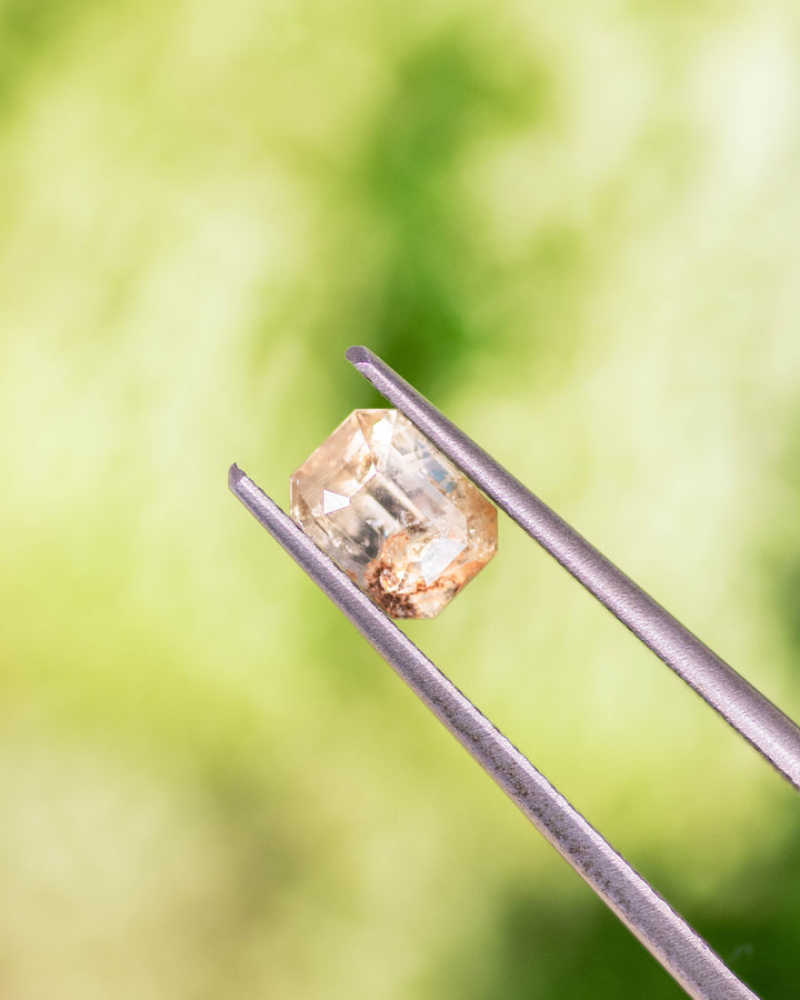 Close-up of 1.11 carat grey, teal, and yellow parti sapphire held in gemstone tweezers, displaying precision step facets and vibrant color zoning of its custom emerald cut, untreated sapphire from Umba River Valley, Tanga, Tanzania.