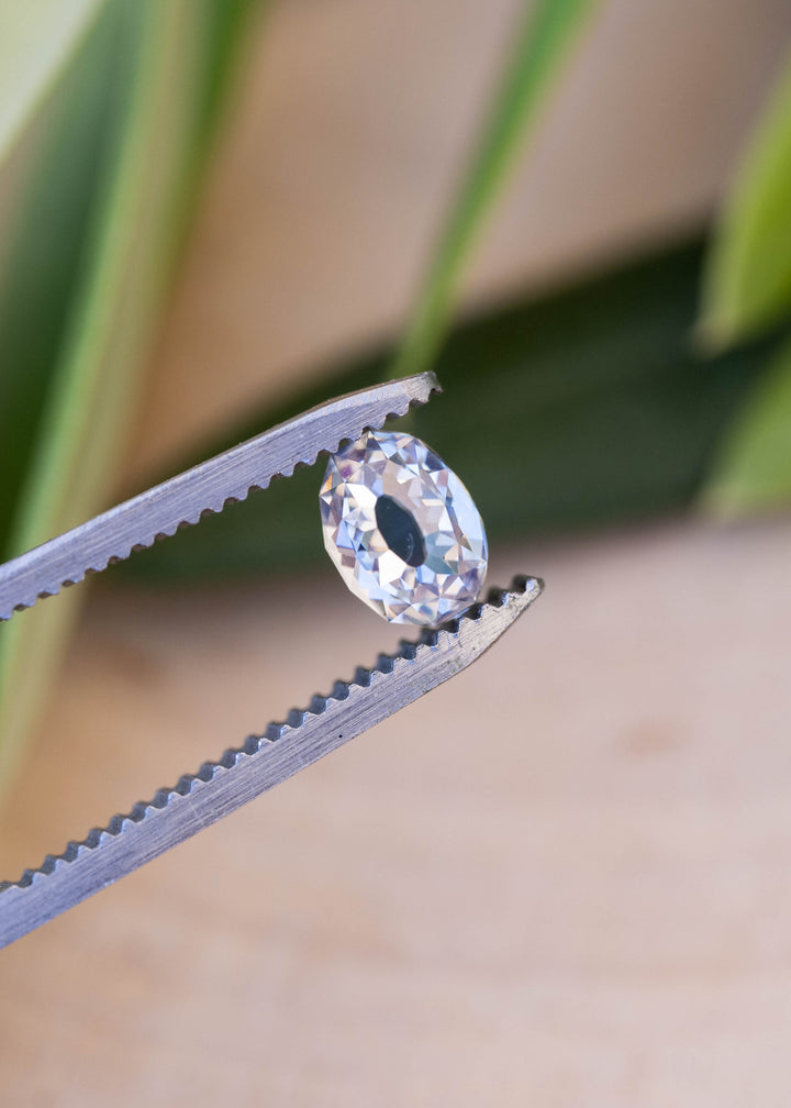 1.11 carat oval diamond held between jeweler’s tweezers against a softly blurred botanical background, showing its elongated outline and open central table.