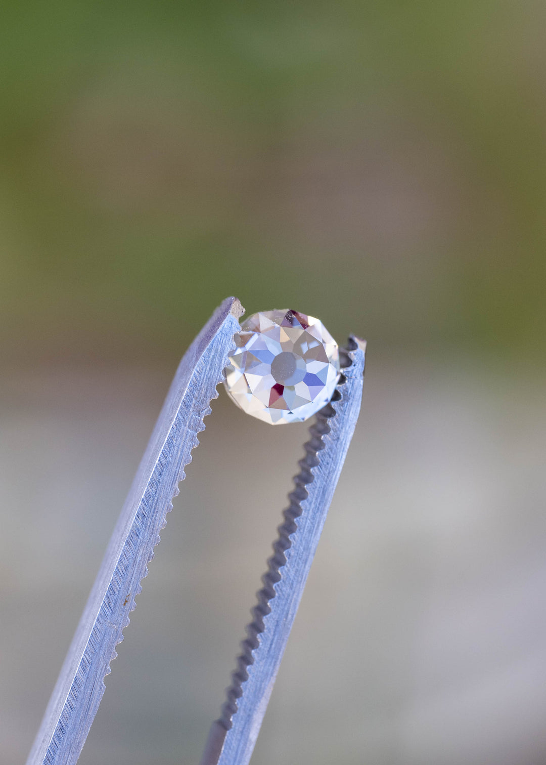 1.11 carat Mughal cut diamond held between jeweler’s tweezers against a softly blurred botanical background, showing its near-round shape and antique-style facet pattern.